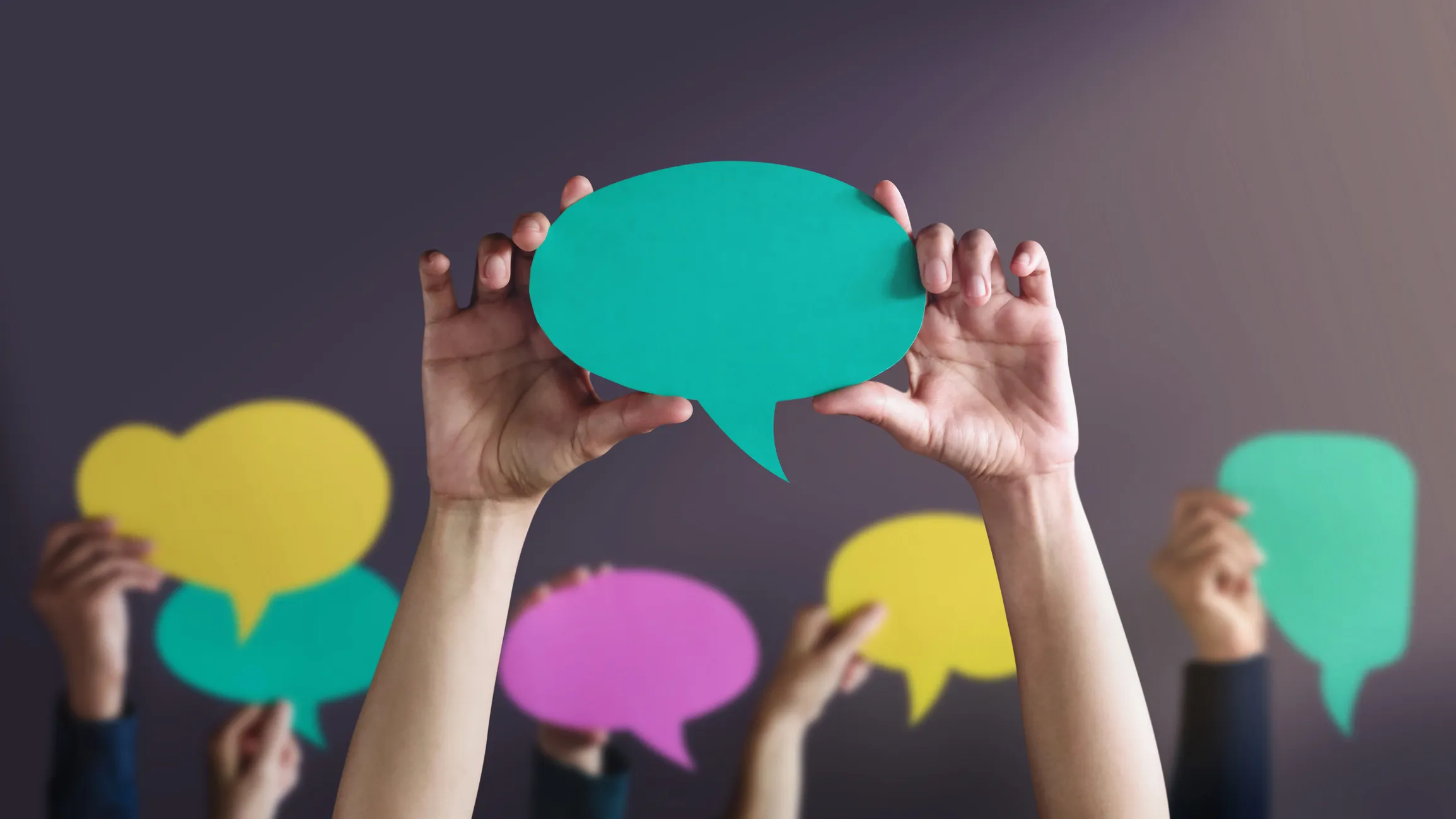 Multiple hands hold up colourful paper speech‑bubble shapes, including teal, yellow, pink, and green, against a dark background to represent communication.