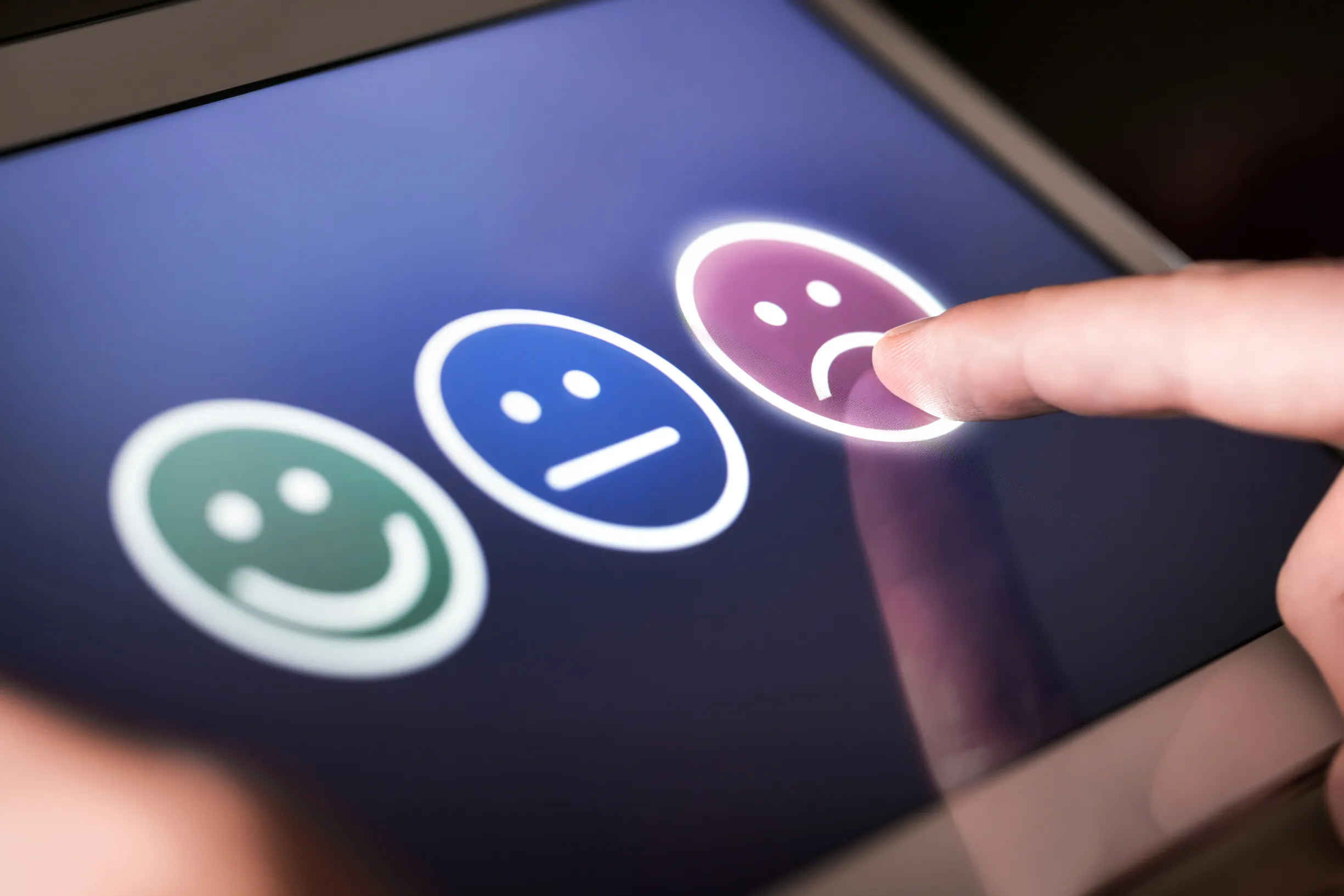 A close-up of a hand touching a tablet screen displaying three face icons: a green smiling face, a yellow neutral face, and a red frowning face. The hand is selecting the red frowning face.